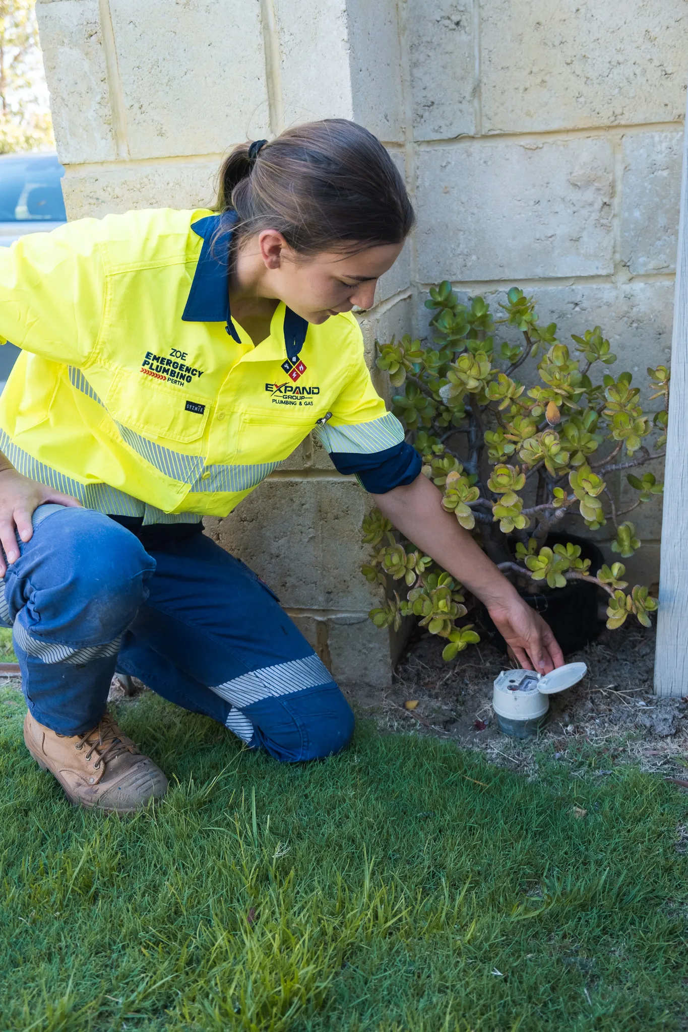 A woman in a high-visibility shirt crouches by a plant, inspecting a device.
